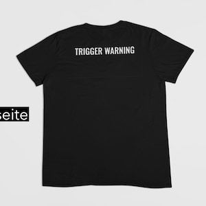Könnte beinhalten: Schwarzes T-Shirt mit dem weißen Aufdruck "TRIGGER WARNING" auf dem oberen Rücken. Das Shirt wird vor einem hellgrauen Hintergrund gezeigt. Das Wort "Rückseite" befindet sich in einem schwarzen Rechteck unten links.