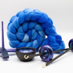 May include: A coiled bundle of blue wool roving sits behind a purple support stand, a small black bowl with a metallic interior, and two drop spindles featuring dark shafts and iridescent purple whorls with small gold-colored metal hooks.