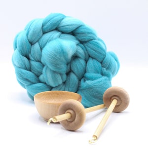 May include: A large, coiled bundle of bright turquoise wool roving positioned behind a small, light-colored wooden bowl and two wooden drop spindles, each featuring a circular whorl and a metal hook at the tip of the shaft.