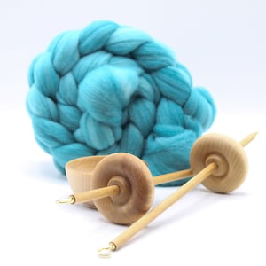 May include: A wooden spinning wheel with two whorls and a spindle. The wheel is made of light brown wood and has a smooth finish. The spindle is made of a darker wood and has a pointed tip. A ball of light blue wool is in the background.