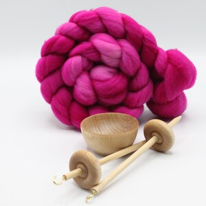 May include: A coiled bundle of vibrant hot pink wool roving positioned behind a small, light-colored wooden bowl and two wooden drop spindles, each featuring a metal hook at the tip and a circular whorl on a slender shaft for spinning fiber.