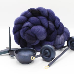 May include: A large, coiled bundle of deep purple and dark blue wool roving positioned behind two dark wooden drop spindles and a small, round dark wooden bowl with a gold-colored metal hook attached to the rim on a plain white background.