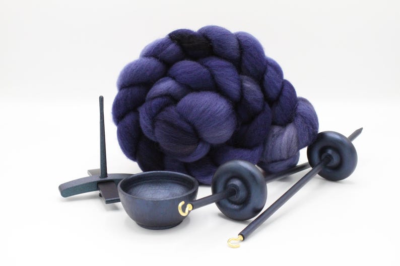 May include: A large, coiled bundle of deep purple and dark blue wool roving positioned behind two dark wooden drop spindles and a small, round dark wooden bowl with a gold-colored metal hook attached to the rim on a plain white background.