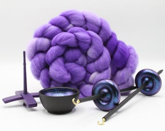 Puede incluir: Un manojo de lana cardada morada, un soporte morado, un cuenco pequeño negro con interior azul y dos husos con ejes negros y torteras azul-moradas iridiscentes. Los husos cuentan con ganchos de color dorado en los extremos.