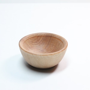 May include: A small, round wooden bowl with a light brown color. The bowl has a smooth, polished finish and a simple, minimalist design. The wood grain is visible on the interior and exterior of the bowl.