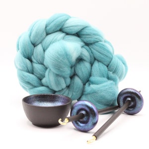 May include: A large coil of teal wool roving positioned behind a small black bowl with a shimmering purple interior and two drop spindles featuring dark shafts and iridescent purple whorls with gold-colored metal hooks at the tips.
