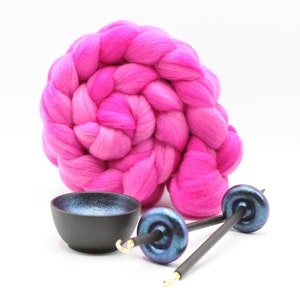 May include: A vibrant hot pink coiled wool roving top positioned behind a small black bowl with a shimmering blue interior and two drop spindles featuring dark, iridescent whorls and thin black shafts with small gold-colored metal hooks.