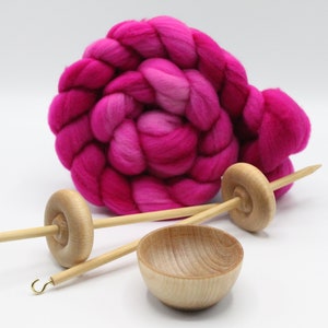 May include: A coiled braid of vibrant hot pink wool roving positioned behind two light-colored wooden drop spindles with metal hooks and a small, matching wooden bowl. The wool features varying shades of magenta and bright pink throughout the fiber.