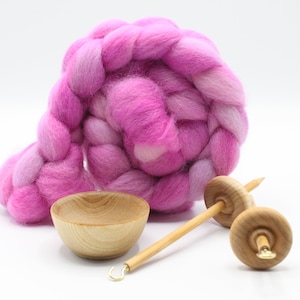 May include: A coiled braid of vibrant pink and light purple wool roving fiber, accompanied by a small light-colored wooden bowl and two wooden drop spindles with metal hooks, all arranged together on a plain white background.