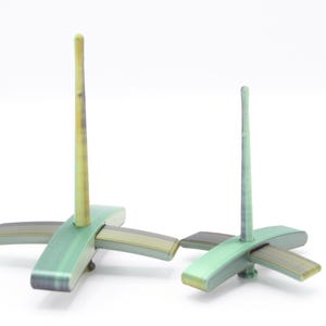 May include: Two abstract sculptures with a cross-shaped base and a vertical rod. The sculptures are painted in shades of green, gray, and yellow. The larger sculpture has a taller rod. The background is white.