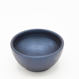 May include: A small, round bowl with a dark, matte navy blue finish. The interior surface features visible, circular brushstroke textures, while the exterior has a consistent, solid dark blue color and a smooth, rounded rim.