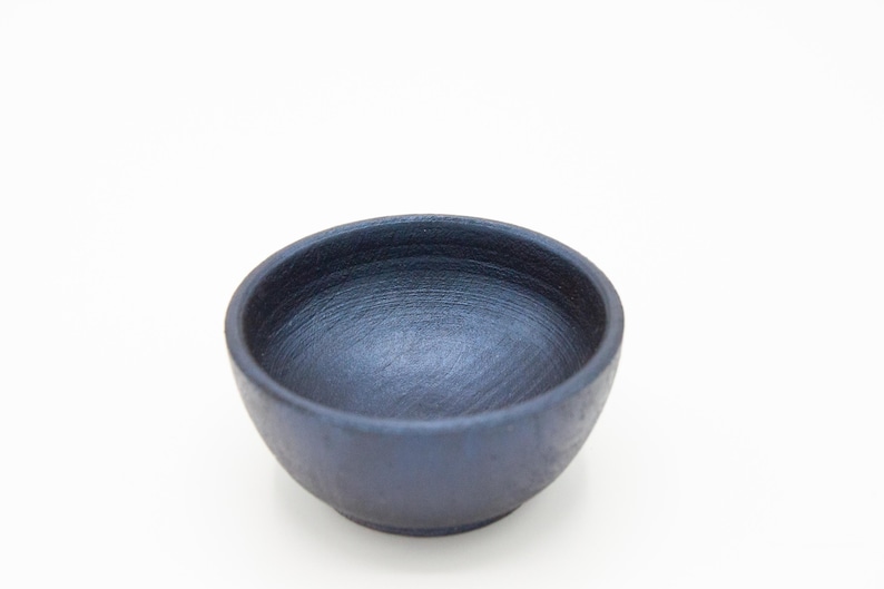 May include: A small, round bowl with a dark, matte navy blue finish. The interior surface features visible, circular brushstroke textures, while the exterior has a consistent, solid dark blue color and a smooth, rounded rim.