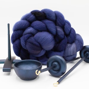 May include: A large, braided bundle of deep blue and purple wool roving positioned behind a dark blue wooden drop spindle, a small matching bowl, and a vertical spindle support stand, all featuring a smooth, matte finish and gold-toned metal hooks.