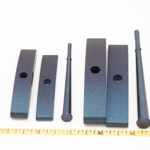 May include: A set of six dark blue metallic tools including four rectangular blocks with central hexagonal holes and two tapered cylindrical rods, arranged horizontally above a yellow measuring tape showing five inches in total length.