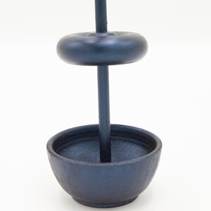 May include: A dark charcoal gray incense burner featuring a vertical central rod, a circular base bowl, and a single removable donut-shaped weight component that slides onto the rod, all constructed with a matte, textured finish.