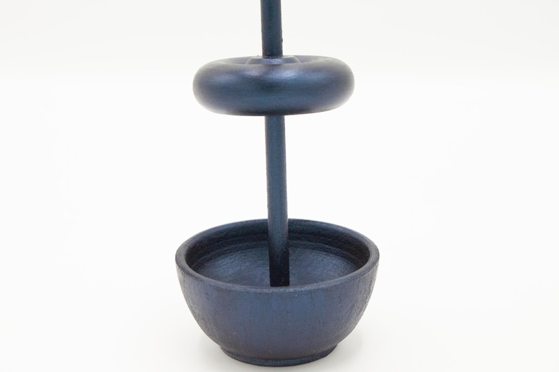 May include: A dark charcoal gray incense burner featuring a vertical central rod, a circular base bowl, and a single removable donut-shaped weight component that slides onto the rod, all constructed with a matte, textured finish.
