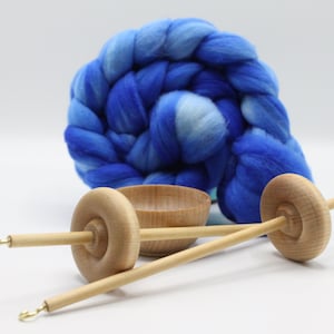 May include: Two light-colored wooden drop spindles with metal hooks at the tips, a small wooden bowl, and a large, thick coil of roving wool fiber featuring a gradient of blue shades ranging from pale sky blue to deep royal blue.