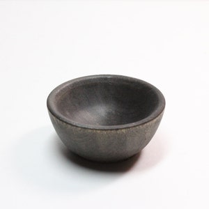 May include: A small, round bowl with a dark charcoal gray finish and a matte, textured surface. The bowl features a thick, rounded rim and a deep, concave interior, showcasing a consistent dark tone throughout its exterior and inner basin.