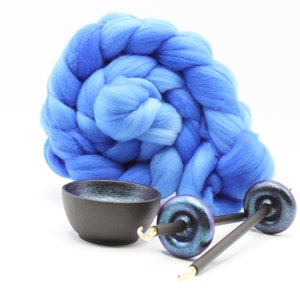 May include: A large, coiled bundle of vibrant blue wool roving positioned behind a small, matte black ceramic bowl and two drop spindles. The spindles feature dark, iridescent whorls with metallic gold-colored hooks at the pointed tips.