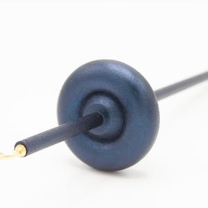 May include: A drop spindle for spinning fiber, featuring a dark blue, circular wooden whorl attached to a long, slender, dark blue wooden shaft. The tip of the shaft includes a small, metallic gold-colored hook for securing yarn during use.