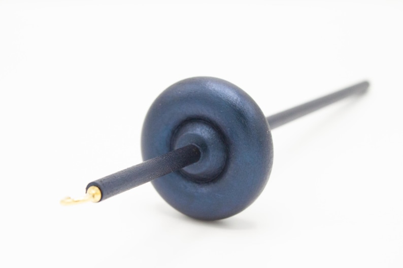 May include: A drop spindle for spinning fiber, featuring a dark blue, circular wooden whorl attached to a long, slender, dark blue wooden shaft. The tip of the shaft includes a small, metallic gold-colored hook for securing yarn during use.