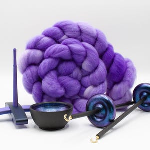 May include: A coiled bundle of purple and violet wool roving positioned behind a purple wooden yarn support, a small black bowl with a blue interior, and two drop spindles featuring dark iridescent whorls and thin metal hooks at the tips.