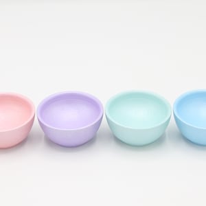 May include: Four pastel-colored bowls in pink, purple, mint green, and light blue are arranged in a row. The bowls are smooth and have a rounded shape, suitable for serving or decorative purposes.