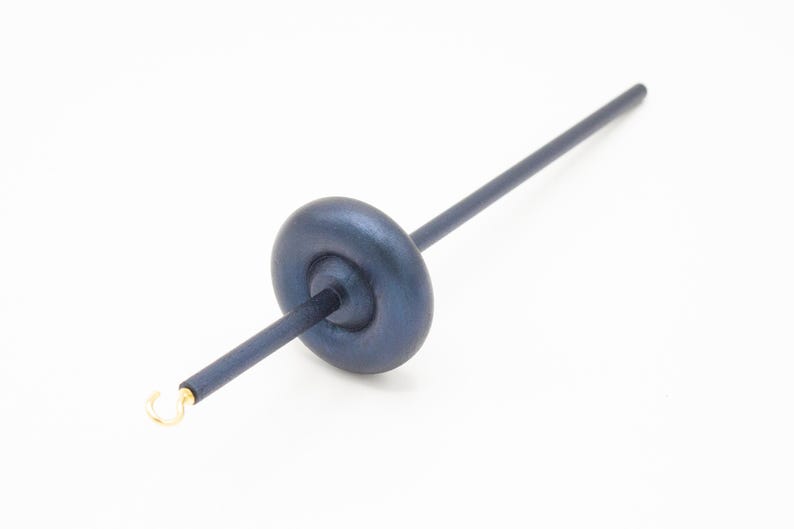 May include: A dark blue wooden drop spindle featuring a smooth, rounded whorl positioned along a straight shaft. The tool includes a small, metallic gold-colored hook at one end, designed for fiber spinning tasks with a sleek, uniform finish.