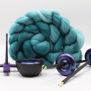 May include: A large coil of teal and turquoise wool roving positioned behind a purple stand, a small black bowl with a glittery interior, and two drop spindles featuring black shafts and iridescent purple whorls with gold-colored hooks.