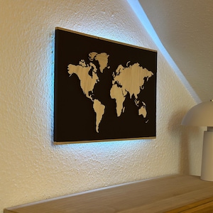 Könnte beinhalten: Eine Weltkarte aus Holz mit hellem Holzfinish ist auf einem schwarzen Hintergrund montiert. Die Karte wird von einem blauen LED-Licht beleuchtet. Eine weiße Lampe mit pilzförmigem Schirm steht auf einer Holzoberfläche.
