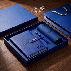 Puede incluir: Un juego de regalo azul que incluye un cuaderno, un bolígrafo y un termo, todos con las iniciales "RM". El juego se presenta en una caja azul a juego, con una bolsa de regalo y clips.