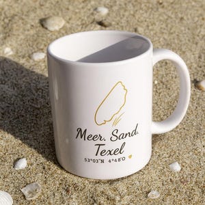Könnte beinhalten: Weiße Keramik-Tasse mit goldener Strandkontur und dem Text "Meer. Sand. Texel" mit Koordinaten. Die Tasse steht auf Sand, umgeben von Muscheln.