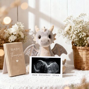 Könnte beinhalten: Ein kuscheliges graues Drachenspielzeug mit großen Augen und Flügeln sitzt neben einem gerahmten Ultraschallbild und einem Buch mit der Aufschrift "Baby Thompson". Auf dem Ultraschallbild steht "Due October 20xx". Die Szene ist mit weißen Blumen dekoriert.