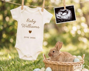 May include: A cream-colored baby onesie with the text "Baby Williams" and a heart, hanging with a sonogram image. A brown rabbit sits in a wicker basket with pastel eggs on green grass.