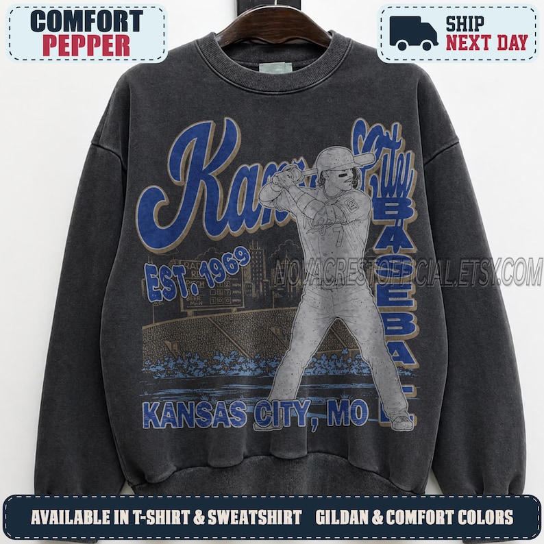 May include: A charcoal gray crewneck sweatshirt featuring a vintage-style graphic of a baseball player at bat. The design includes blue text reading Kansas City Baseball, Est. 1969, and Kansas City, MO, set against a stadium background illustration.