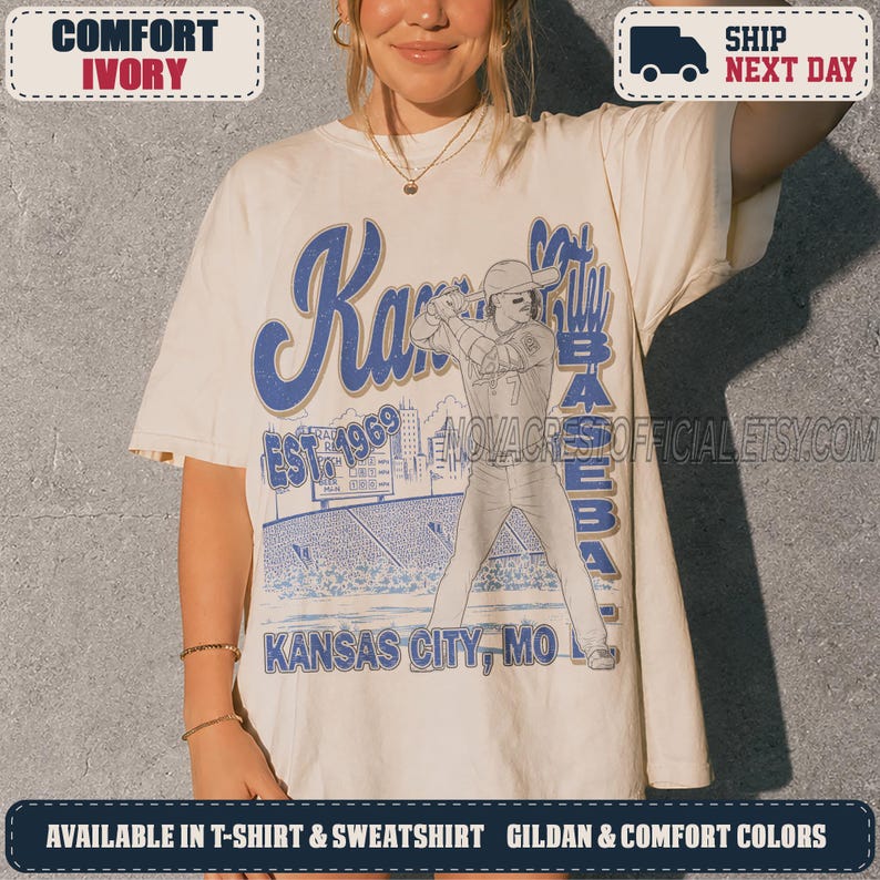 May include: An ivory t-shirt featuring a blue graphic print of a baseball player at bat. The text reads Kansas City Baseball, Est. 1969, and Kansas City, MO. The design includes a stadium background and a scoreboard, rendered in a vintage style.