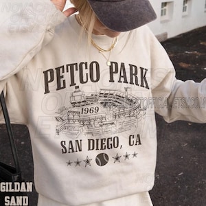 May include: A sand-colored sweatshirt with the text "PETCO PARK 1969 SAN DIEGO, CA" in black. The design includes a baseball stadium illustration and a baseball graphic. The sweatshirt is a Gildan brand.