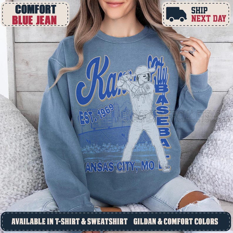 May include: A blue crewneck sweatshirt featuring a graphic of a baseball player at bat. The text reads Kansas City Baseball Est. 1969 Kansas City, MO. The design includes a stadium illustration in blue and white tones on a solid blue fabric background.