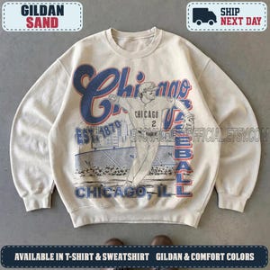 May include: A sand-colored crewneck sweatshirt featuring a large graphic print of a baseball player in a pitching stance. The design includes the text Chicago Baseball, Est. 1870, and Chicago, Ill, with a stadium background in blue and red ink.