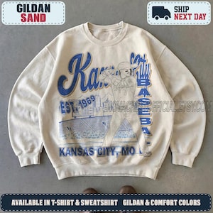 May include: A sand-colored crewneck sweatshirt featuring a blue graphic print of a baseball player at bat. The design includes the text Kansas City Baseball, Est. 1969, and Kansas City, MO, along with a stadium illustration and scoreboard.