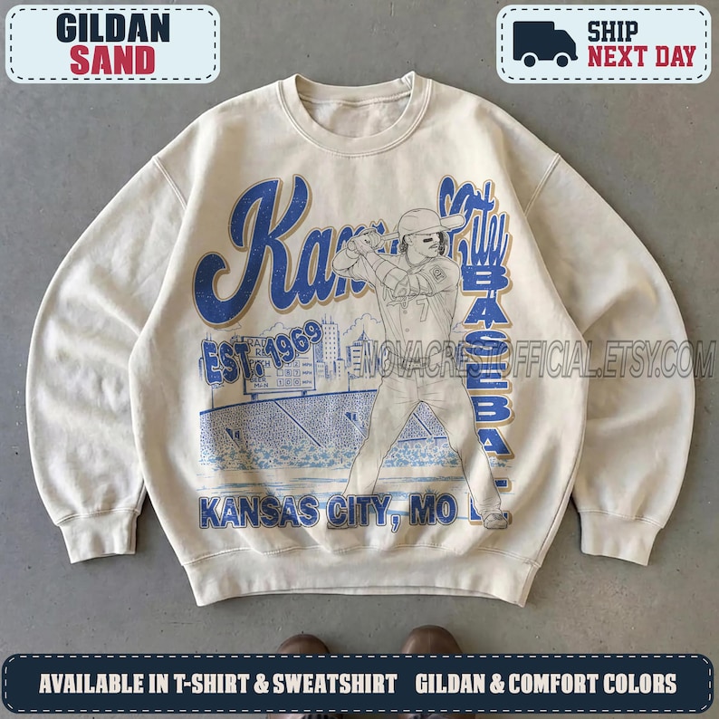 May include: A sand-colored crewneck sweatshirt featuring a blue graphic print of a baseball player at bat. The design includes the text Kansas City Baseball, Est. 1969, and Kansas City, MO, along with a stadium illustration and scoreboard.