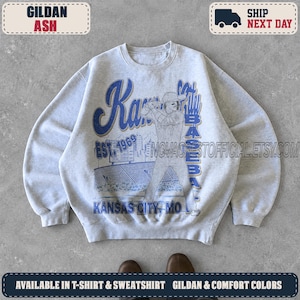 May include: A light gray crewneck sweatshirt featuring a blue graphic print of a baseball player at bat in a stadium. The text reads Kansas City Baseball, Est 1969, and Kansas City, MO. The design includes a scoreboard and stadium seating.