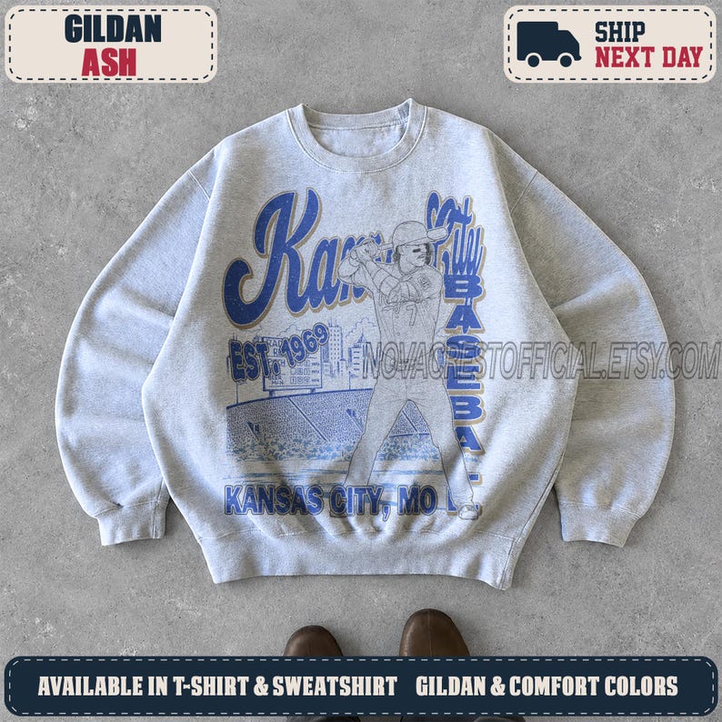 May include: A light gray crewneck sweatshirt featuring a blue graphic print of a baseball player at bat in a stadium. The text reads Kansas City Baseball, Est 1969, and Kansas City, MO. The design includes a scoreboard and stadium seating.