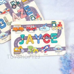 May include: Wooden puzzle with the name "JAYCE" in colourful letters and vehicle shapes, including a school bus, fire engine, and rocket. The puzzle also features a road design and the word "UFO".