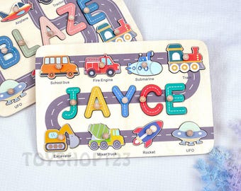 Op de afbeelding: Houten puzzel met de naam "JAYCE" in kleurrijke letters en voertuigvormen, waaronder een schoolbus, brandweerwagen en raket. De puzzel heeft ook een wegontwerp en het woord "UFO".