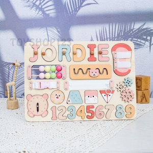 K&ouml;nnte beinhalten: Holz-Lernspielzeug mit dem Namen "JORDIE" in bunten Buchstaben. Das Brett enth&auml;lt verschiedene interaktive Elemente, darunter einen Rei&szlig;verschluss, Perlen, Zahnr&auml;der und Zahlen. Eine Uhr und Tierformen sind ebenfalls enthalten.