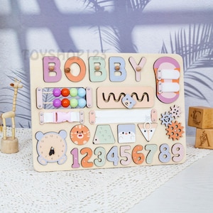 K&ouml;nnte beinhalten: Holz-Lernspielbrett mit dem Namen "BOBBY" in bunten Buchstaben. Enth&auml;lt eine Uhr, Zahlen, Formen, Zahnr&auml;der, einen Rei&szlig;verschluss und Perlen. Ein unterhaltsames und ansprechendes Spielzeug f&uuml;r Kleinkinder.