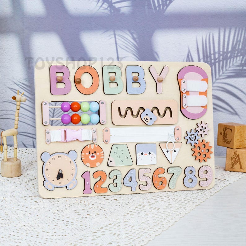 K&ouml;nnte beinhalten: Holz-Lernspielbrett mit dem Namen "BOBBY" in bunten Buchstaben. Enth&auml;lt eine Uhr, Zahlen, Formen, Zahnr&auml;der, einen Rei&szlig;verschluss und Perlen. Ein unterhaltsames und ansprechendes Spielzeug f&uuml;r Kleinkinder.