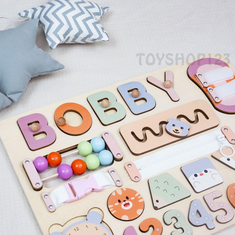 K&ouml;nnte beinhalten: Holz-Lernspielzeug mit dem Namen "BOBBY" in bunten Buchstaben. Zu den Merkmalen geh&ouml;ren ein Perlen-Abakus, ein Rei&szlig;verschluss, eine Schnalle sowie verschiedene Formen und Zahlen. Enth&auml;lt ein Katzen- und B&auml;r-Design.