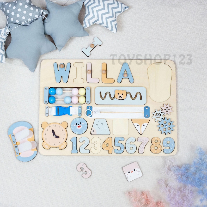 K&ouml;nnte beinhalten: Holz-Lernspielzeug mit dem Namen "WILLA" in blauen und orangenen Buchstaben. Das Brett enth&auml;lt verschiedene interaktive Elemente, darunter ein Abakus, Rei&szlig;verschluss, Schnalle, Uhr und Zahlen. Der Text "TOYSHOP123" ist sichtbar.